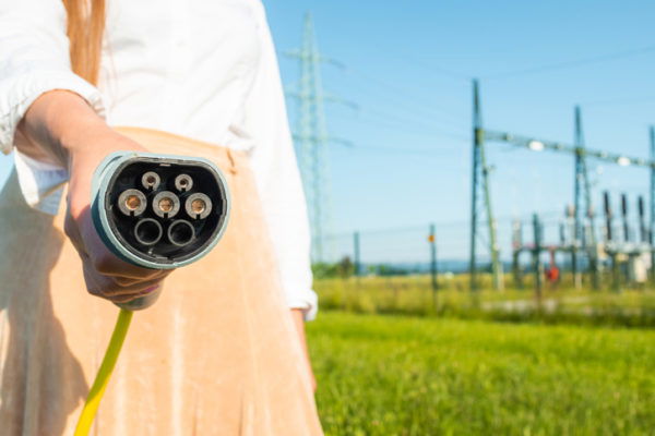 Woman unplugging electric car charger at a charging station near electric power plant, in the background visible transmission line