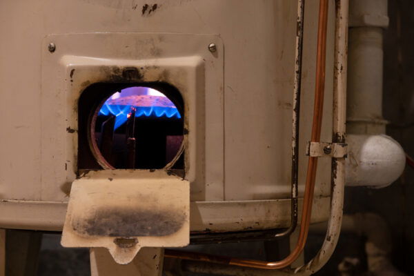 Fragment of an old white gas heating boiler. There are metal fittings and pipes. An open window shows a blue flame on the burner
