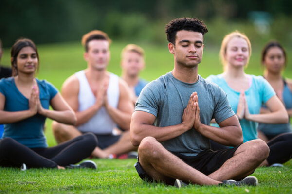 A group of multi-ethnic young adults sit cross-legged outside on the grass together on a sunny summer day.