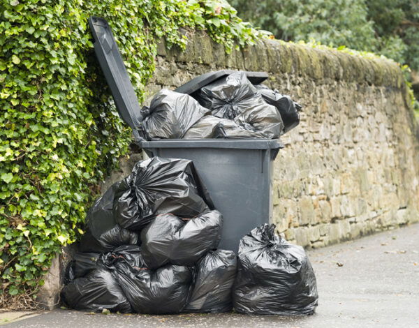 A full wheelie bin with rubbish bags overflowing on to the pavement