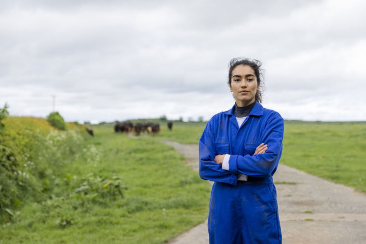 A portrait of a young female farmer wearing overalls, standing in an agricultural field at the farm she works at in Embleton, North East England while looking at the camera with a serious look on her face and her arms crossed